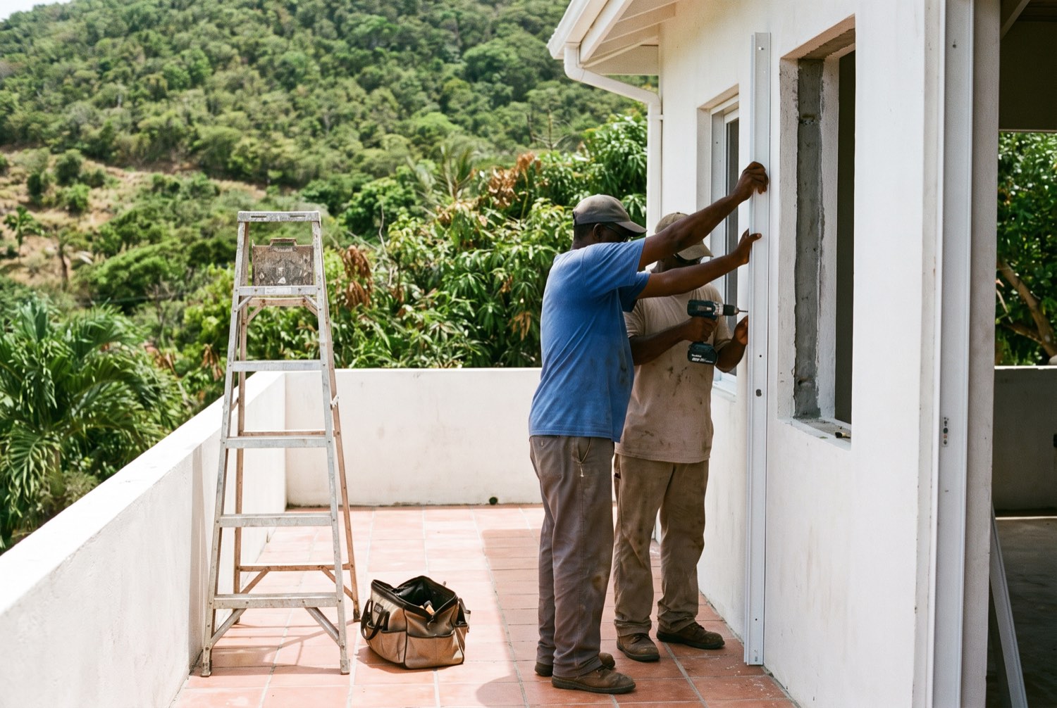 Two tradesmen fitting a white aluminium guide rail to a window reveal on a Caribbean house terrace, a step ladder and tool bag beside them.