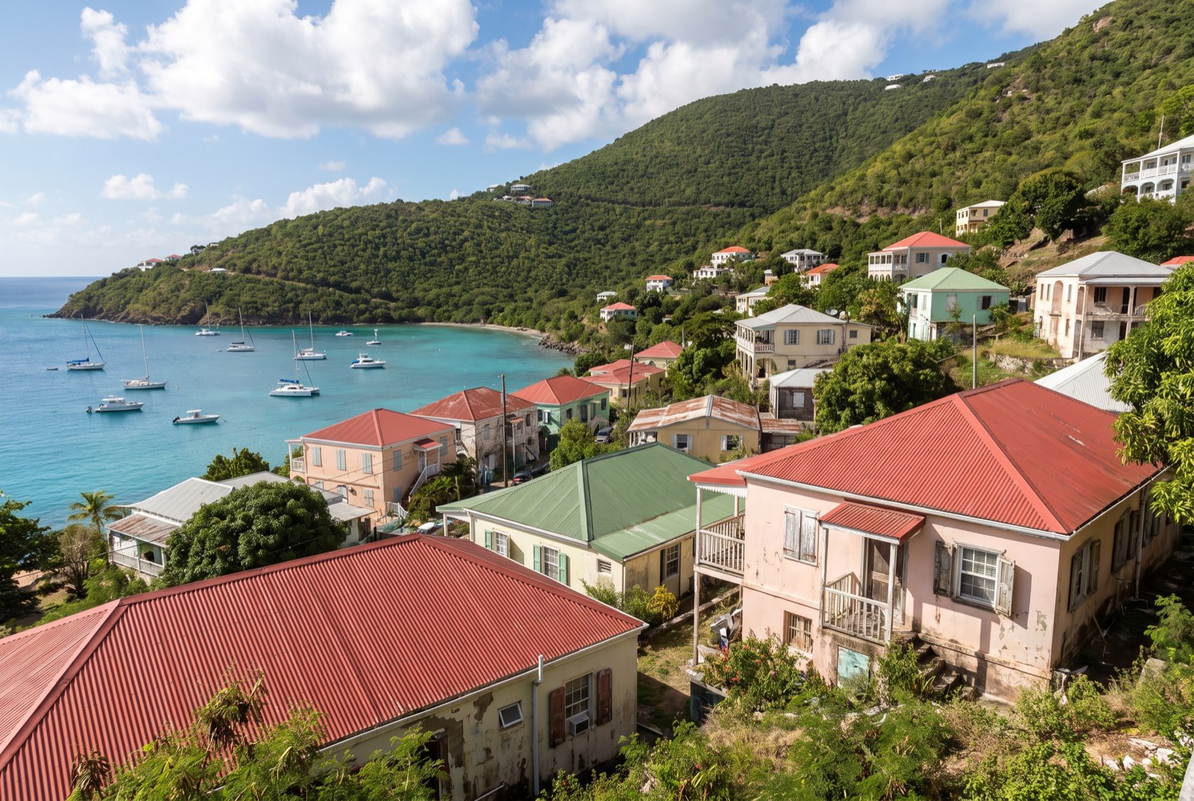 A Virgin Islands hillside of white and pastel houses with red galvanized roofs stepping down toward a turquoise bay.